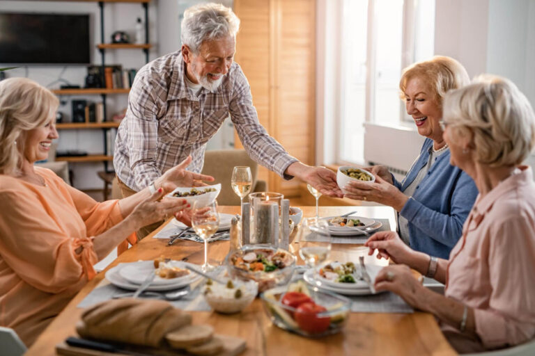 Grupo de amigos idosos sorrindo e compartilhando uma refeição. Volte a comer seus alimentos favoritos com a prótese protocolo em Cruz Alta.